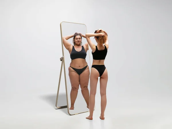 Молодая стройная женщина, смотрящая на толстую девушку в зеркалах, отражающихся на белом фоне — стоковое фото