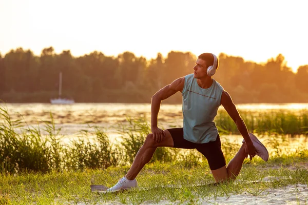 Un joven atlético haciendo ejercicio escuchando la música a la orilla del río al aire libre — Foto de Stock