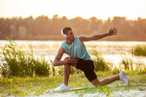 Un joven atlético haciendo ejercicio escuchando la música a la orilla del río al aire libre — Foto de Stock