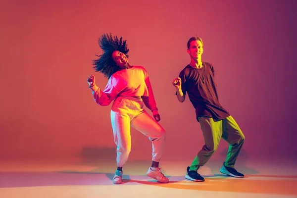 Stylish man and woman dancing hip-hop in bright clothes on gradient background at dance hall in neon light