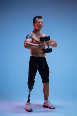 Özürlü ya da yaralı bir sporcu mavi stüdyo arka planında izole edilmiş. Profesyonel erkek sporcu, bacak protezi eğitimi almış, ağırlıkları neon.