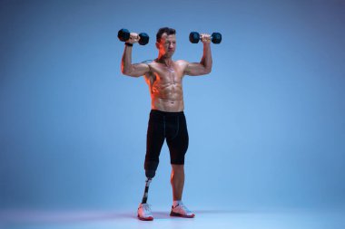 Özürlü ya da yaralı bir sporcu mavi stüdyo arka planında izole edilmiş. Profesyonel erkek sporcu, bacak protezi eğitimi almış, ağırlıkları neon.