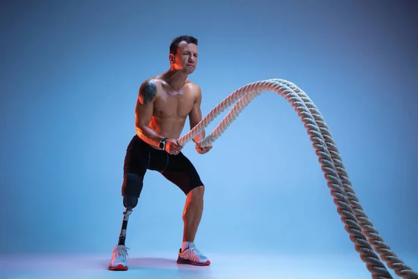 Athlete with disabilities or amputee isolated on blue studio background. Professional male sportsman with leg prosthesis training with weights in neon