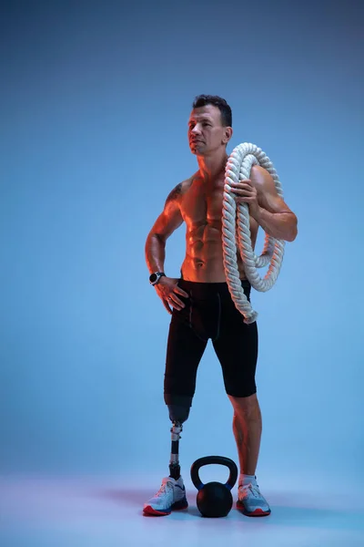 Atleta con discapacidad o amputado aislado en el fondo del estudio azul. Deportista masculino profesional con entrenamiento de prótesis de pierna con pesas en neón — Foto de Stock