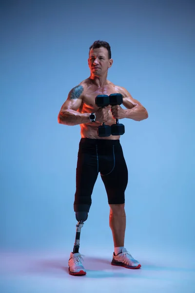 Atleta con discapacidad o amputado aislado en el fondo del estudio azul. Deportista masculino profesional con entrenamiento de prótesis de pierna con pesas en neón — Foto de Stock