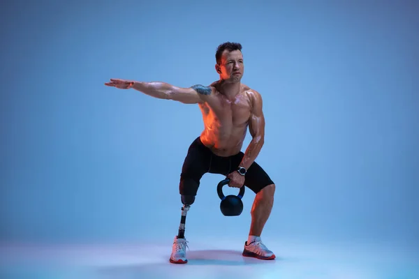 Athlete with disabilities or amputee isolated on blue studio background. Professional male sportsman with leg prosthesis training with weights in neon