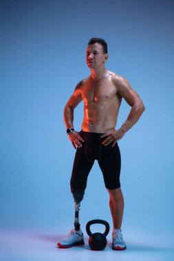 Özürlü ya da yaralı bir sporcu mavi stüdyo arka planında izole edilmiş. Profesyonel erkek sporcu, bacak protezi eğitimi almış, ağırlıkları neon.