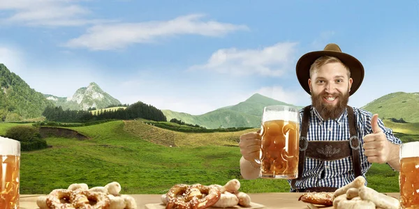 El hombre sonriente feliz con cerveza vestida con traje tradicional austriaco o bávaro sosteniendo taza de cerveza, montañas en el fondo, volante —  Fotos de Stock