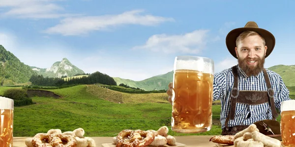 El hombre sonriente feliz con cerveza vestida con traje tradicional austriaco o bávaro sosteniendo taza de cerveza, montañas en el fondo, volante —  Fotos de Stock