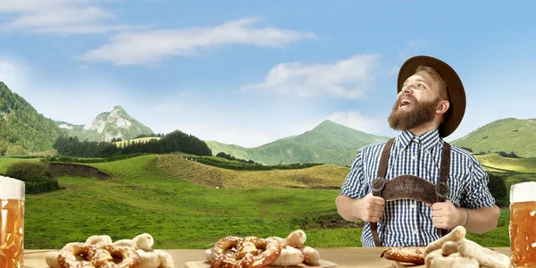 Счастливый улыбающийся человек с пивом, одетый в традиционный австрийский или баварский костюм, держащий кружку пива, горы на заднем плане, флаер — стоковое фото