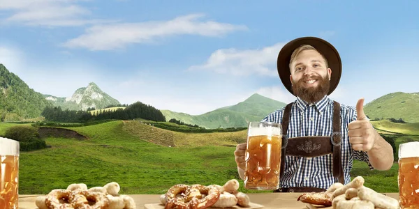 El hombre sonriente feliz con cerveza vestida con traje tradicional austriaco o bávaro sosteniendo taza de cerveza, montañas en el fondo, volante —  Fotos de Stock