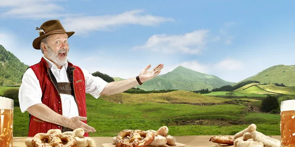 The senior happy smiling man with beer dressed in traditional Austrian or Bavarian costume holding mug of beer, mountains on background, flyer — Stock Photo, Image