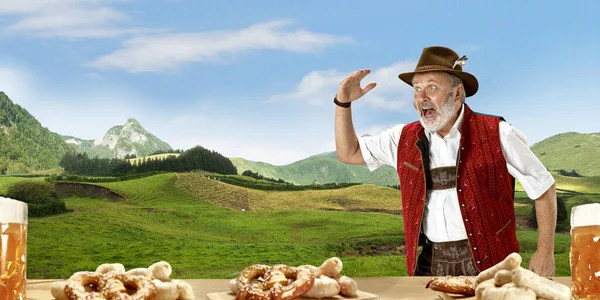 The senior happy smiling man with beer dressed in traditional Austrian or Bavarian costume holding mug of beer, mountains on background, flyer — Stock Photo, Image