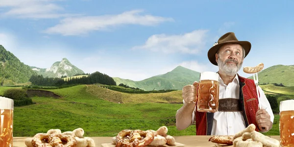 The senior happy smiling man with beer dressed in traditional Austrian or Bavarian costume holding mug of beer, mountains on background, flyer — Stock Photo, Image