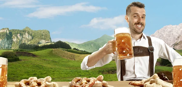 El hombre sonriente feliz con cerveza vestida con traje tradicional austriaco o bávaro sosteniendo taza de cerveza, montañas en el fondo, volante —  Fotos de Stock