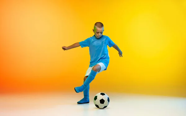 Mladý chlapec jako fotbalista nebo fotbalista ve sportovním oblečení cvičení na gradient žluté pozadí studia v neonovém světle — Stock fotografie