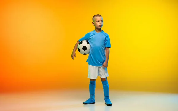 Young boy as a soccer or football player in sportwear practicing on gradient yellow studio background in neon light — Stock Photo, Image