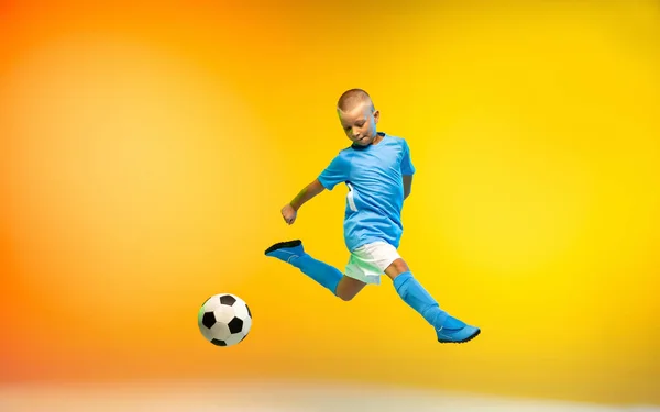 Young boy as a soccer or football player in sportwear practicing on gradient yellow studio background in neon light