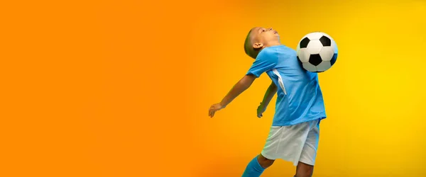 Young boy as a soccer or football player in sportwear practicing on gradient yellow studio background in neon light — Stock Photo, Image