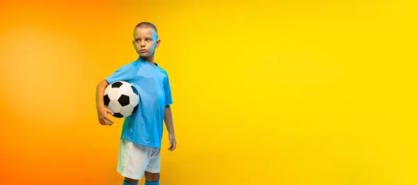 Jeune garçon comme footballeur ou joueur de football en tenue de sport pratiquant sur fond de studio jaune dégradé au néon — Photo