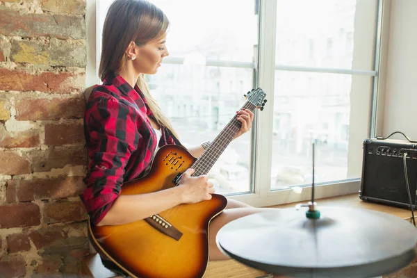 Mujer grabando música, cantando y tocando la guitarra mientras está sentada en el lugar de trabajo o en casa — Foto de Stock