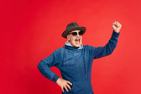 Homem sênior em óculos elegantes e chapéu isolado no fundo vermelho. Conceito de tecnologia e estilo de vida idoso alegre — Fotografia de Stock