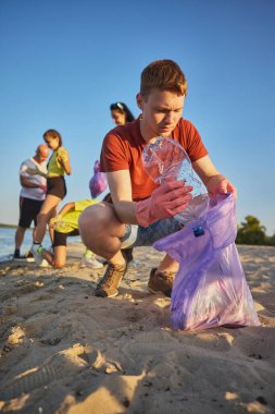 Kırmızı gömlekli genç adam odaklı olarak kumlu sahilde plastik şişe topluyor. Çevreye yönelik çevre sorumluluğu, farkındalık, sürdürülebilirlik ve duygusal bakım kavramı.