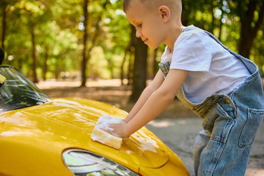 Sarı oyuncak araba kaputunu sünger ve köpükle temizleyen bir çocuk. Sorumluluk kavramı, erken gelişim, açık hava oyunları, olumlu alışkanlıklar ve çocukluk sevinci..