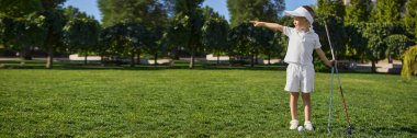 Beyaz üniformalı çocuk golfçü yeşil alanda golf sopalarıyla işaret ediyor. Açık hava sporları, çocukluk eğitimi, kendine güven, odaklanma ve sağlıklı yaşam tarzı kavramı. Pankart