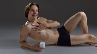 Rahatlamış genç adam göğsüne krem sürerken gülümsüyor. Cilt bakımı kampanyaları, erkek refahı markalaşması, tımar ürünleri ve yaşam tarzı reklamcılığı kavramı.