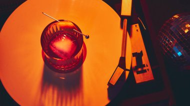 Buzlu kokteyl bardağı ve retro ışıkta parlayan vinil pikabın üzerine garnitür. Gece hayatı pazarlama, alkol markalaşması, duygusal atmosfer ve yaratıcı sanat fotoğrafçılığı kavramı..