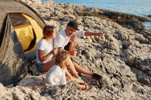 Side view of friendly family sitting near tent at rock beach, admiring sea.