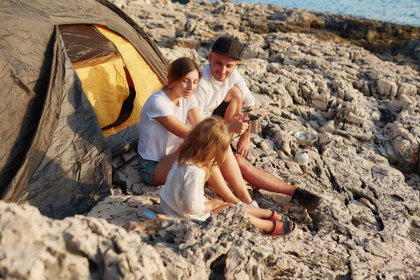Amable familia joven, sentado en el suelo cerca de la tienda de campaña en la costa de piedra . —  Fotos de Stock