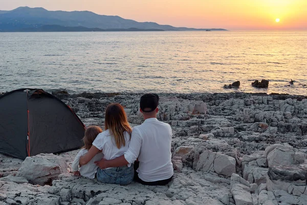 Achteraanzicht van familie, zittend op een rots strand, u geniet van de ondergaande zon over zee. — Stockfoto