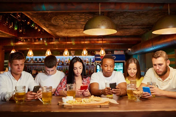 Group of six friends with serious facial expression, sitting with smartphones, drinking beer at bar.