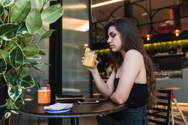 Güler yüzlü lezzetli taze bardak tutan ve şık kafe masada oturan rahat kıyafet içinde çekici genç kadın yan görünüm