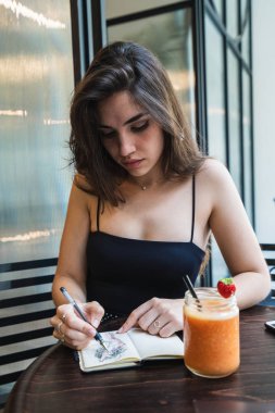 Güler yüzlü lezzetli bardak yakınındaki yuvarlak kafeterya masada oturan ve küçük sketchbook çizim rahat kıyafet çok güzel genç bayan