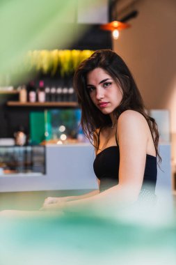 Genç ve güzel esmer kameraya otururken bakarak yan görünümü arka plan bulanık bar