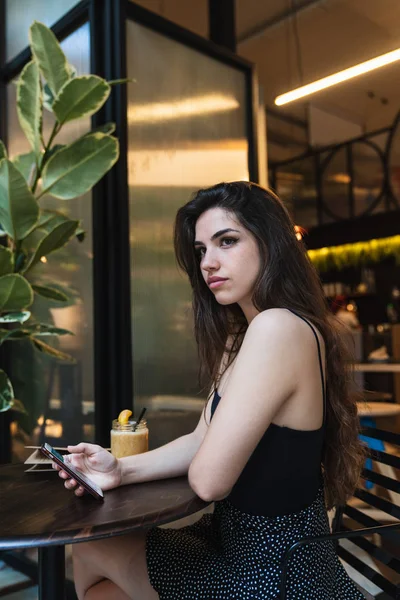 Smartphone holding ve uzak görünümlü şık kafede oturan ve rahatlatıcı güzel genç esmer yan görünüm