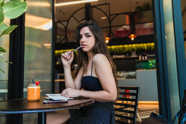 Güzel genç bayan kalem ısırma ve şık kafede oturan ve çizim yaparken kameraya bakarak yan görünüm