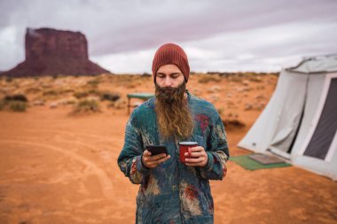 Monument Valley, Utah bulutlu gün çadır uzak olmayan bir yerde dururken modern smartphone kullanarak sıcak içecek fincan ile çekici sakallı adam