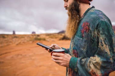 Kırpma yan görünüm Utah bulutlu gün inanılmaz Anıt Vadisi dururken modern smartphone tarama taze sıcak içecek bir fincan adamla sakallı