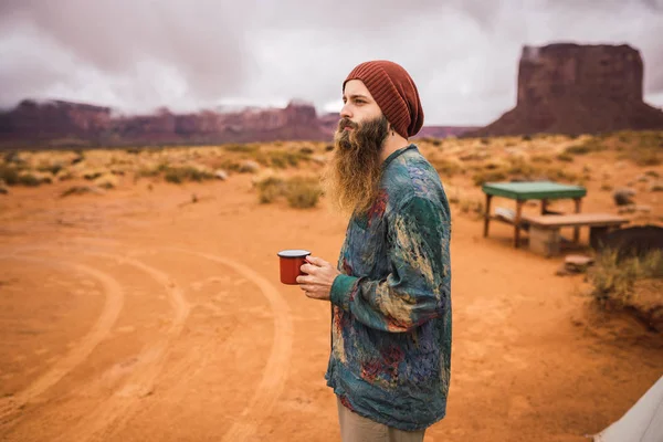 侧面视图的胡子家伙在时尚的衣服持有杯热饮和看着一边站在露营区在阴天纪念碑山谷 犹他州 — 图库照片