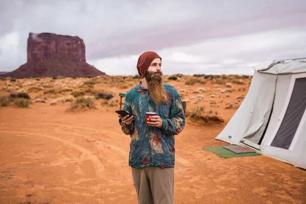 英俊的胡子家伙与杯子的热饮浏览智能手机和看着一边站在纪念碑山谷 犹他州的帐篷附近 — 图库照片