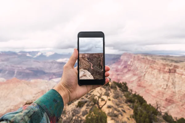 在明亮的衬衫站在山顶的人的手和手持智能手机与大峡谷的图片在亚利桑那州多云天空背景 — 图库照片