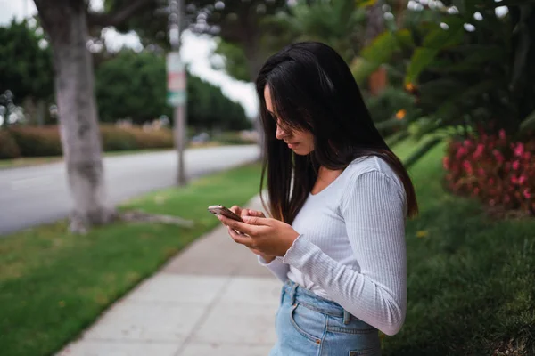 漂亮年轻女性的侧面视图站立在人行道和浏览现代智能手机在洛杉矶 加利福尼亚州 — 图库照片