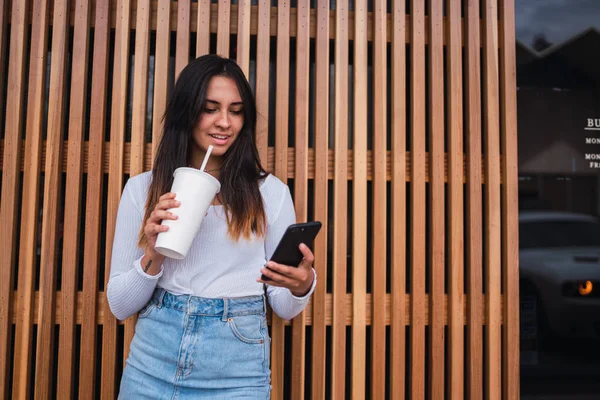 Lovely young female in stylish outfit enjoying tasty beverage and browsing modern smartphone while standing near lumber wall of cafe on street of Los Angeles, California