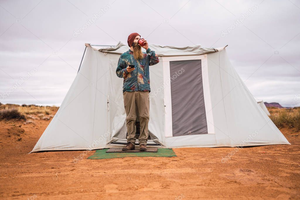 Bearded guy with modern smartphone enjoying hot drink and looking away while standing near big tent in Monument Valley, Utah