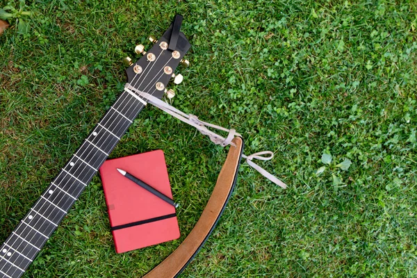 From above shot of small notepad with red cover and pen lying on green grass near guitar