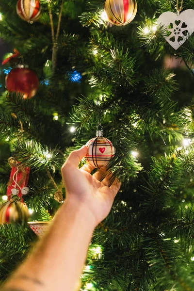 Crop male hand hanging and touching beautiful glass bauble on shiny Christmas tree